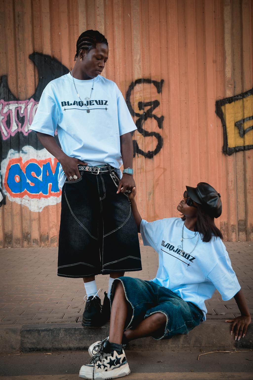 Two adults in trendy streetwear pose by a graffiti wall, exuding urban style.